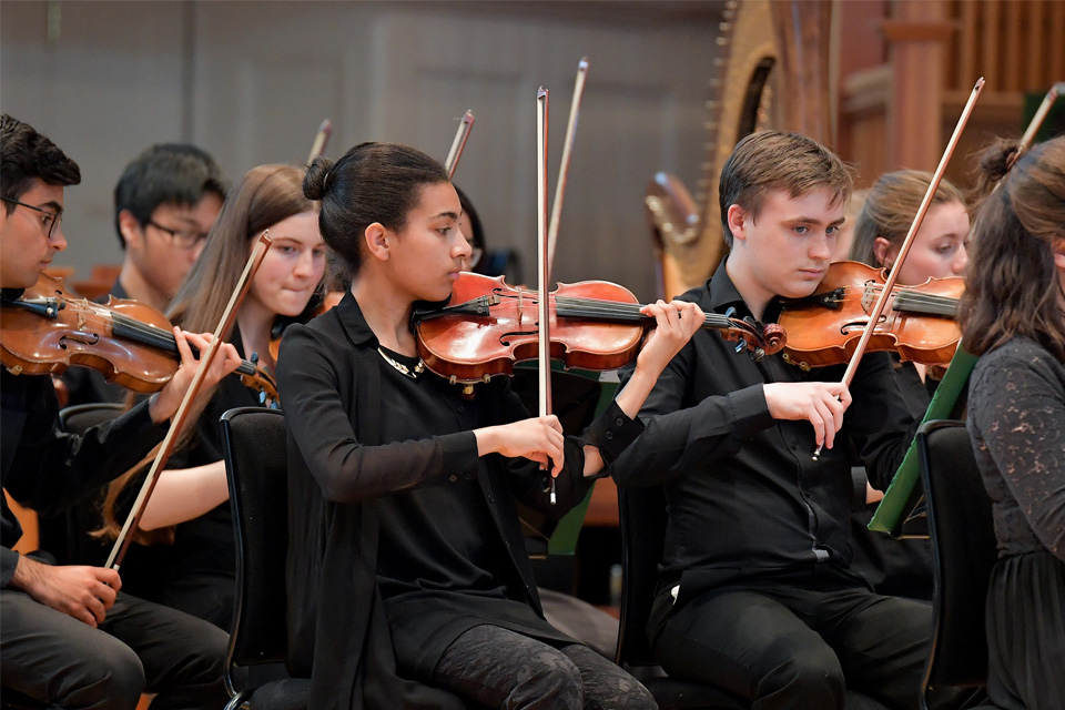 A group of young students, wearing a black smart attire, performing on the violin, in an orchestral performance.